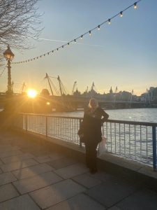 Anita is leaning against a railing with the Thames river behind her and the sun setting to the left of the image
