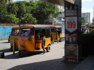 Yellow auto rickshaws, also known as tuk tuks are parked at the end of a road with trees and buildings in the background. A person is getting in or out of one of the tuk tuks and there is a sign advertising chicken buckets on a pillar to the right hand side of the image.