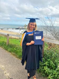 Lauren stands smiling at the camera in a graduation cap and gown, holding her degree certificate. Bournemouth beach is in the background behind her.