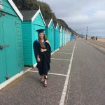 Olivia standing in front of blue beach huts on Bournemouth beach wearing her graduation gown