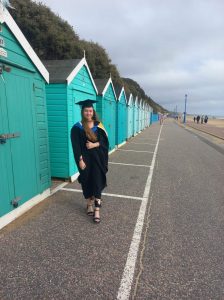 Olivia stands on Bournemouth seafront, wearing a graduation cap and gown with turquoise beach huts behind her
