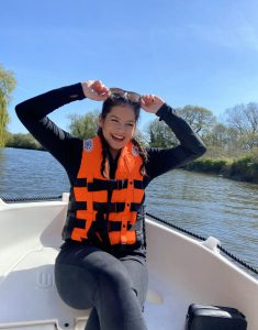 Shanley is sitting in a boat with a life jacket on with water behind her. She is smiling and reaching for sunglasses on top of her head. 
