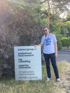 Sam stands next to a sign for Science Group PLC, wearing black jeans, a white tee shirt with a graphic on it and sunglasses