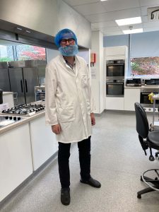 Sam stands in a lab wearing a white lab coat, a hair net and beard net