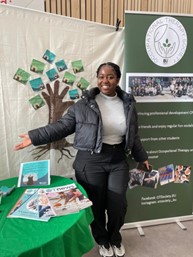 Yaa is smiling and standing in front of a stand and banner for the Occupational Therapy Society