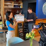 Two people are standing in front of a restaurant bar, and there is a camera facing them. The man is holding a menu looking down at it and the woman is holding the clapperboard