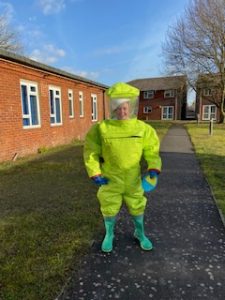 Kylie is wearing a bright green hazmat suit outdoors with blue skies overhead and red brick buildings around her