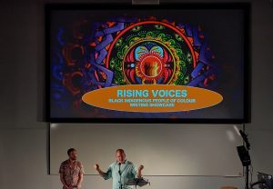 Two people standing in front of a presentation with the words Rising Voices 