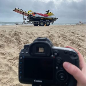 Hannah's hand holds a camera, the backdrop to this is the sandy beach, overcast blue/grey sky and the RNLI's branded jet ski on a transportation vehicle.