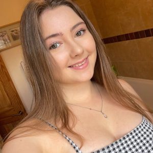 Hannah smiles to the camera in this selfie style image. she wears a strappy black and white checked top and a dainty silver chain necklace. 