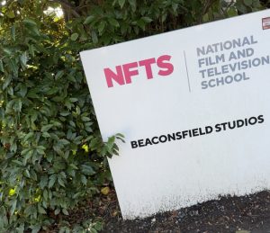 A green leaved bush and a white board sits in front of it with the words NFTS National Film and Television School Beaconsfield Studios 