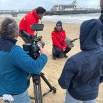the backs of two camera operators, wearing rain macs are shown kneeling on the beach infront of two RNLI staff wearing red rain macs. Bournemouth pier is pictured behind them and a fluffy boom microphone can be seen to the right of the image.