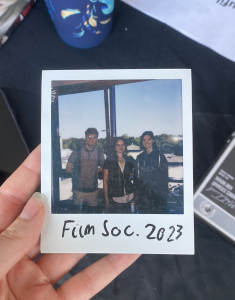 Hand holding polaroid photo of three people- a man and two women- the committee for film society 2023 / 2024. "Film Soc 2023" is written on the polaroid.