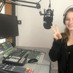 Smiling girl, sitting, holding up her fingers in peace signs, in the Nerve Radio station. There is a mic and radio desk in front of her.