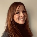 BU Law student, Susie smiles at the camera in a selfie style photo. Her auburn hair is long, draped over her shoulders. She wears a grey roll neck jumper.