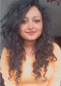 Unnati smiles to the camera, her long curly dark hair surrond her face and falls onto her shoulders. She is pictured in a selfie style photos and wears a light orange top. 