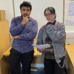 Imran and Rosie perch on a table in a seminar room posing with their thumb and finger of their chin in a comical thought pose. Rosie holds a copy of her Duke of Imran Certificate. 